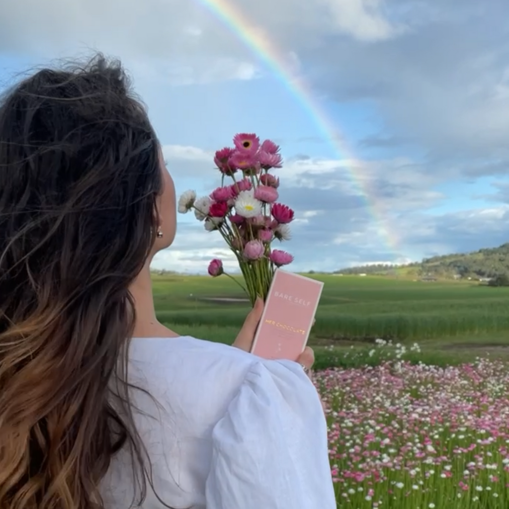 Bare Self, Her Chocolate original, held by girl with flowers, in flower field. rainbow in background. 